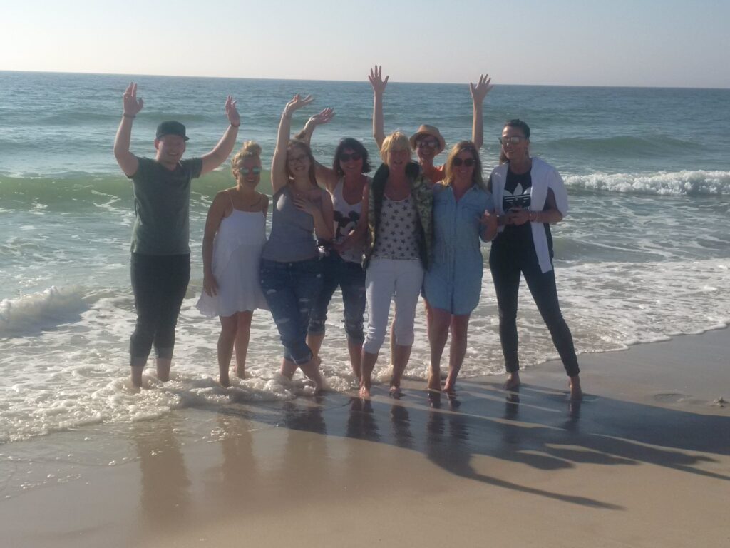 Eine Gruppe von neun Personen steht am Ufer eines Strandes, die Wellen plätschern sanft an ihre Füße. Sie lächeln und winken in die Kamera, im Hintergrund sind das Meer und der klare Himmel zu sehen.