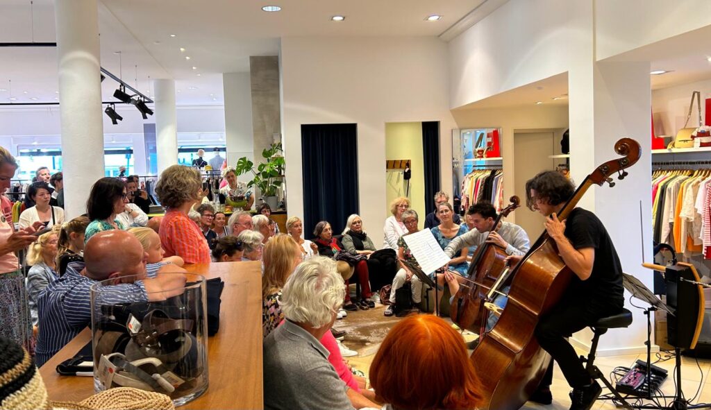 Ein kleines Publikum hat sich in einem Verkaufsraum versammelt und schaut zwei Musikern beim Cellospiel zu. Der Raum ist voller sitzender und stehender Menschen. Im Hintergrund sind Kleiderständer zu sehen. Die Atmosphäre wirkt ungezwungen und engagiert.