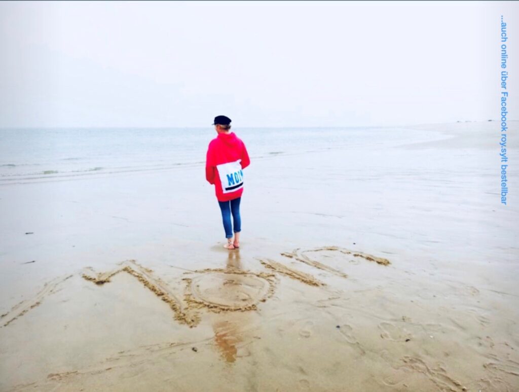 Eine Person mit einem roten Kapuzenpullover und einer schwarzen Kappe steht an einem Sandstrand mit Blick auf das Meer. Vor ihr ist das Wort „Mom“ in den Sand geschrieben. Über ihrer Schulter hängt eine Stofftasche mit dem Text.