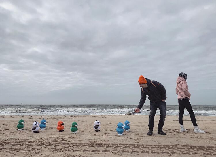 Zwei Menschen stehen an einem wolkenverhangenen Strand. Einer legt bunte Gummienten in den Sand, während der andere zusieht. Im Hintergrund sind die Wellen des Meeres zu sehen.